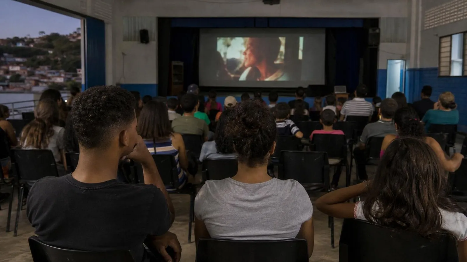 Estudantes e famílias assistem a uma sessão de cinema em um auditório público simples.